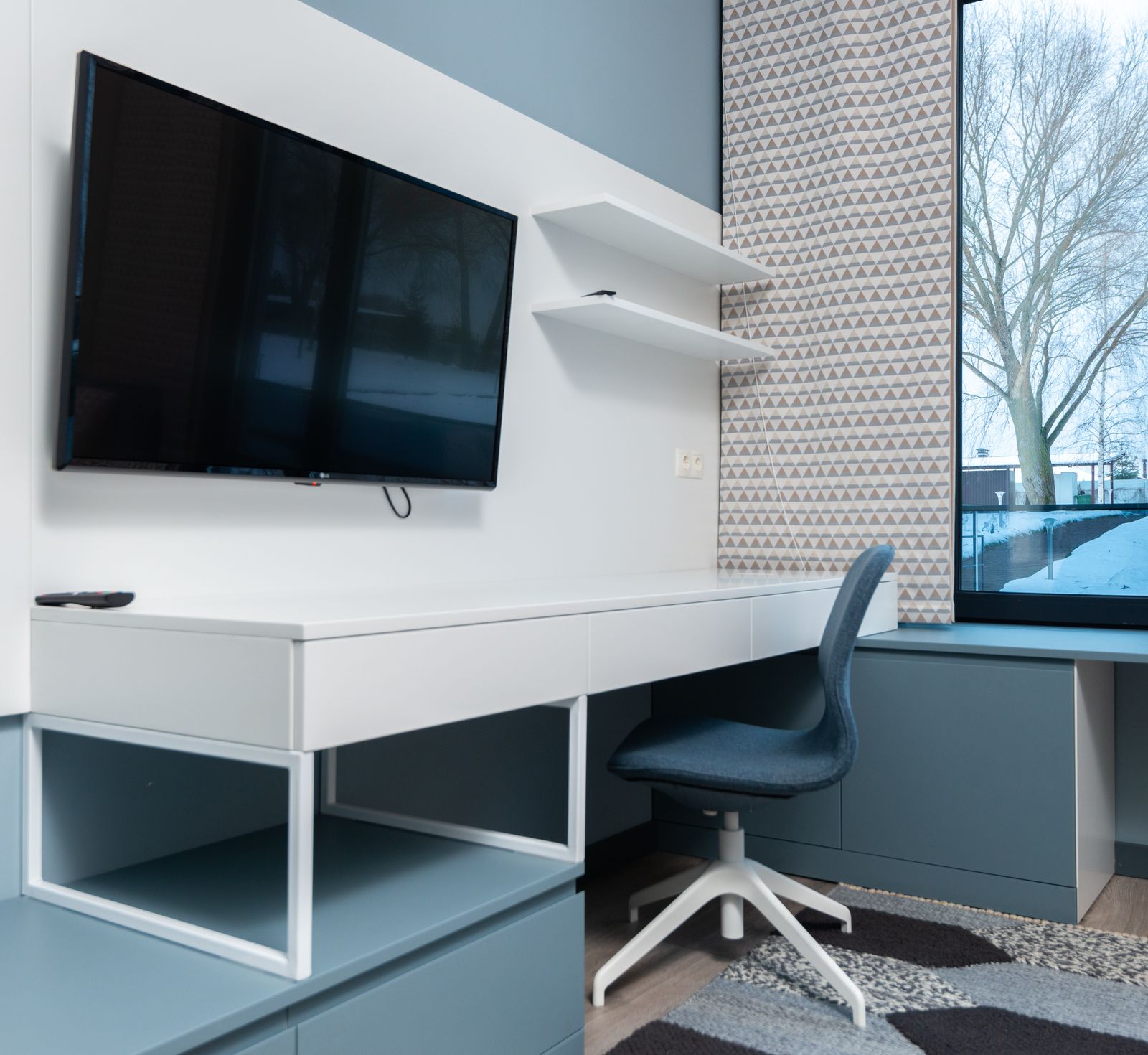 A flat-screen TV mounted on a wall above a simple work table with a chair