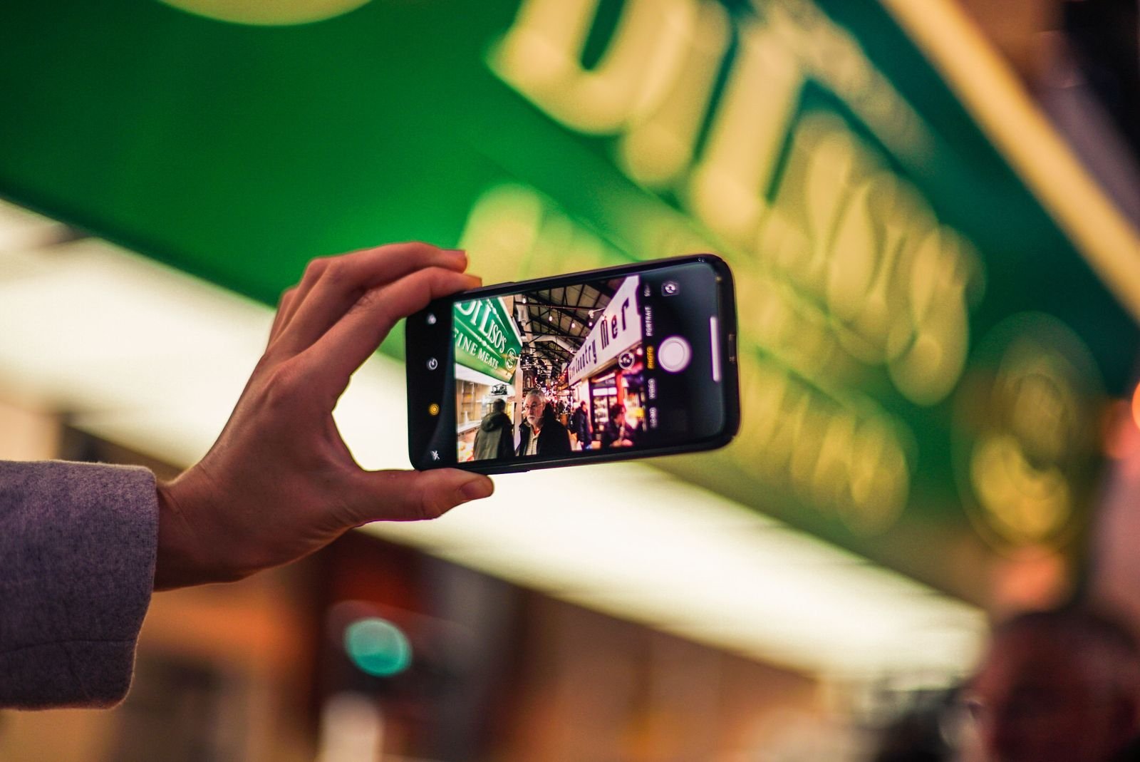 A hand holding a smartphone up to photograph a busy indoor market, with a large green shop sign blurred in the background and the same sign readable on the phone's screen