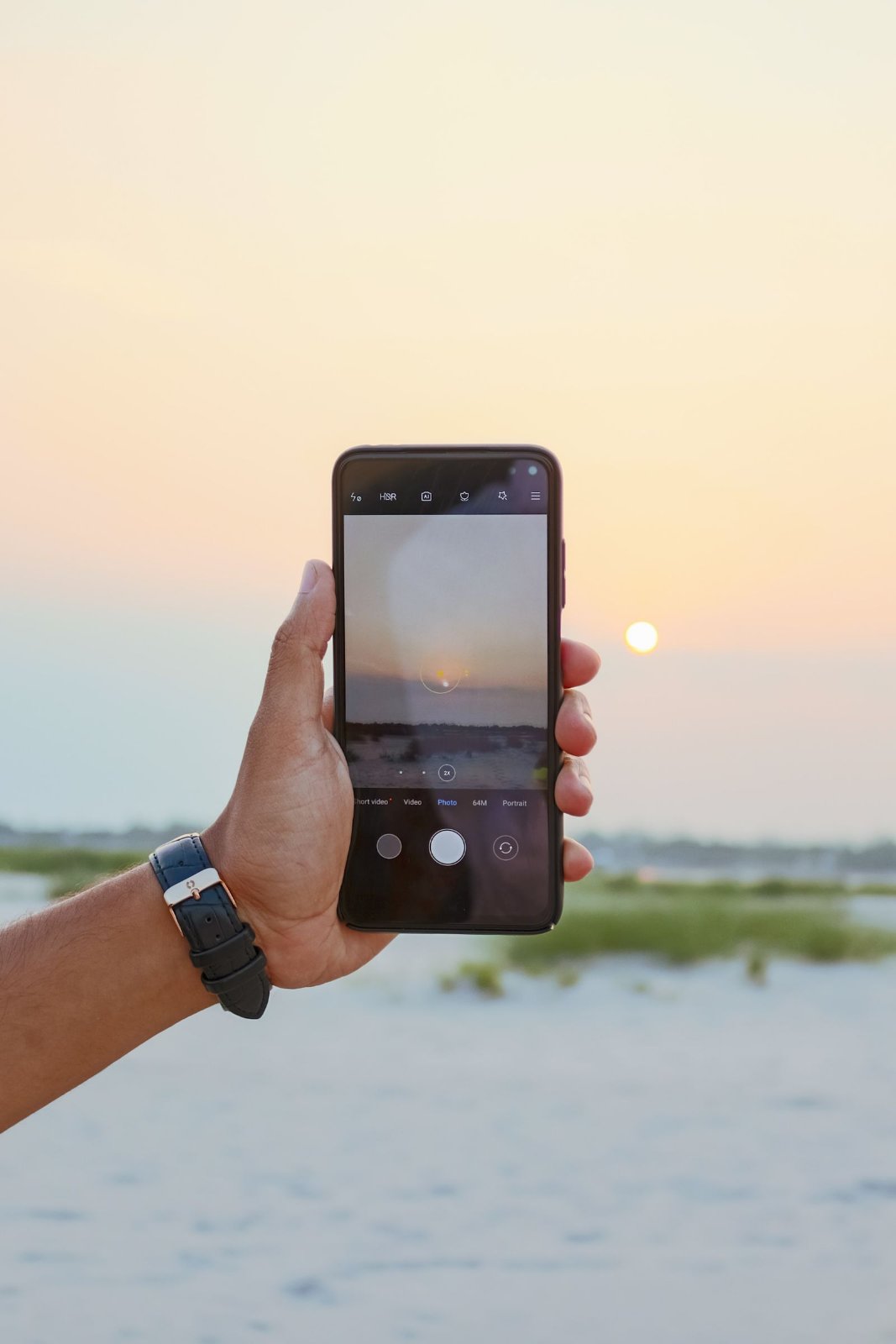 A hand wearing a black leather watch holds up a smartphone at the beach, framing a low sun over a sandy beach on the phone's camera screen