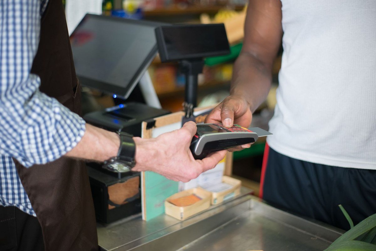 A shop employee holding a handheld card reader while a customer holds a bank card to it at a small grocery store checkout counter