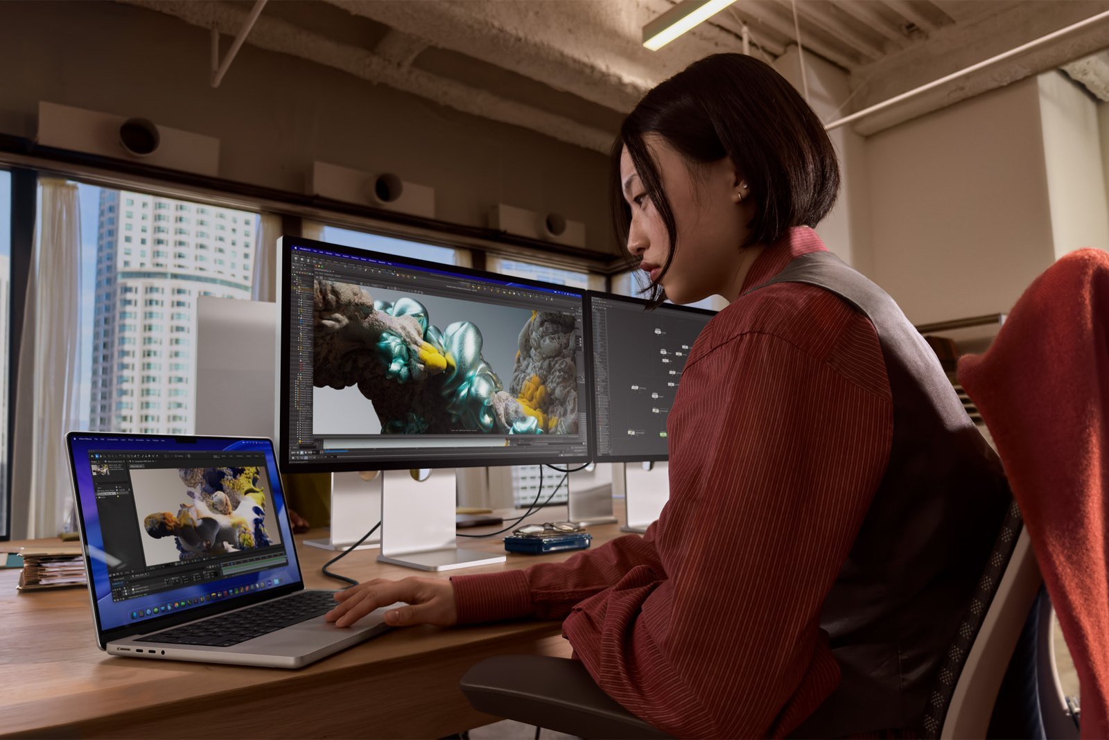 A person at a desk working on a MacBook Pro connected to two external monitors showing 3D rendering software, in a sunlit city office