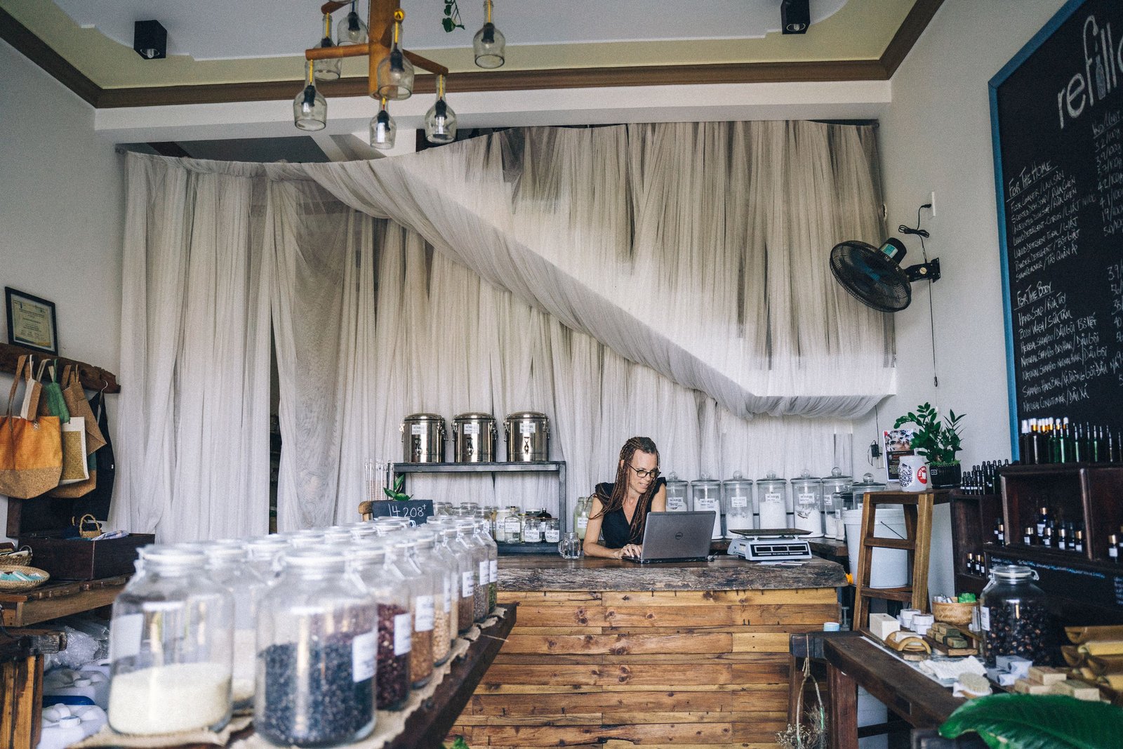 A woman working at a laptop at a shop counter, shelves of stock behind her